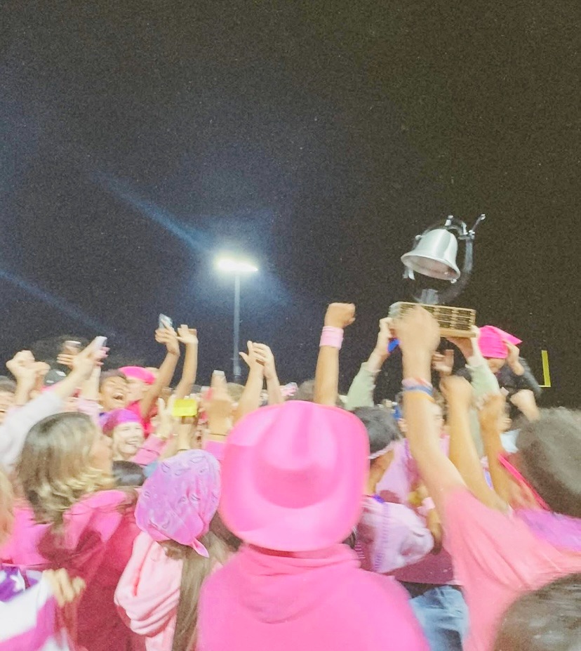 A jubilant crowd of students in pink attire celebrating while holding a trophy aloft during a night event.