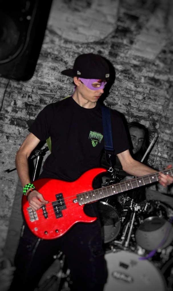 A young musician wearing a purple mask and black cap plays a red bass guitar on stage, with a drummer visible in the background.