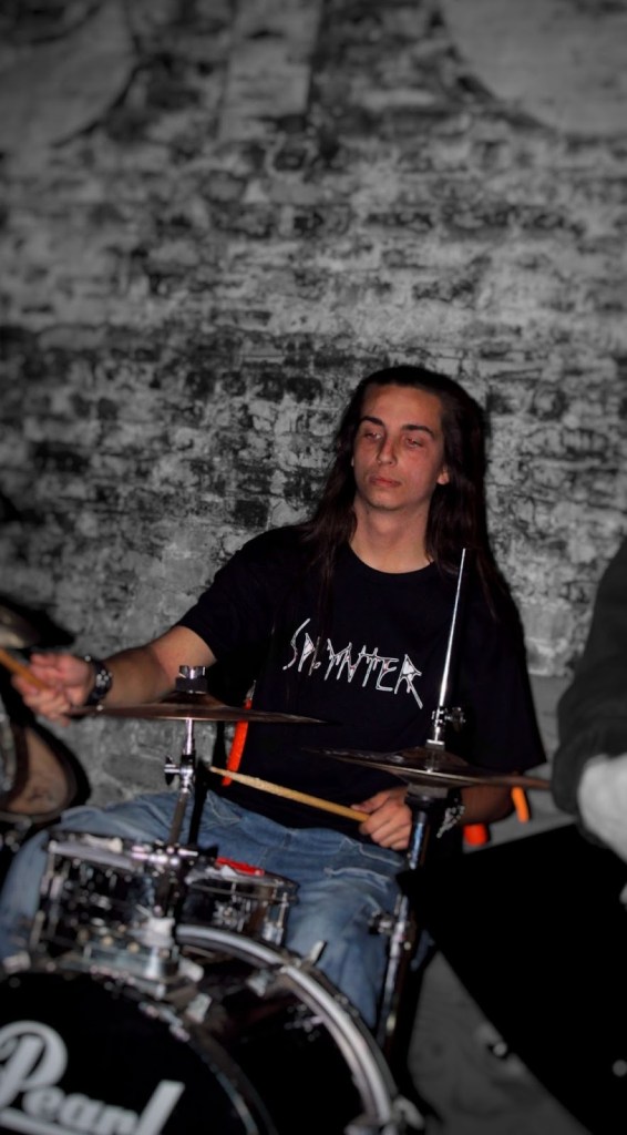 A drummer playing in a dimly lit venue, wearing a black t-shirt with the band name 'SPLYNTER' printed on it. The backdrop features exposed brick walls.