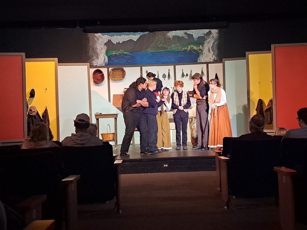 A scene from the UHS Drama Department's production of Babette's Feast, featuring a group of actors in period costumes rehearsing on stage in a theater setting.