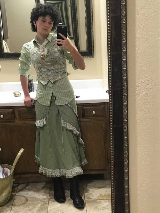A student poses in a stylish outfit featuring a layered green ensemble, complete with a patterned vest, long skirt, and black combat boots, while taking a selfie in front of a mirror.