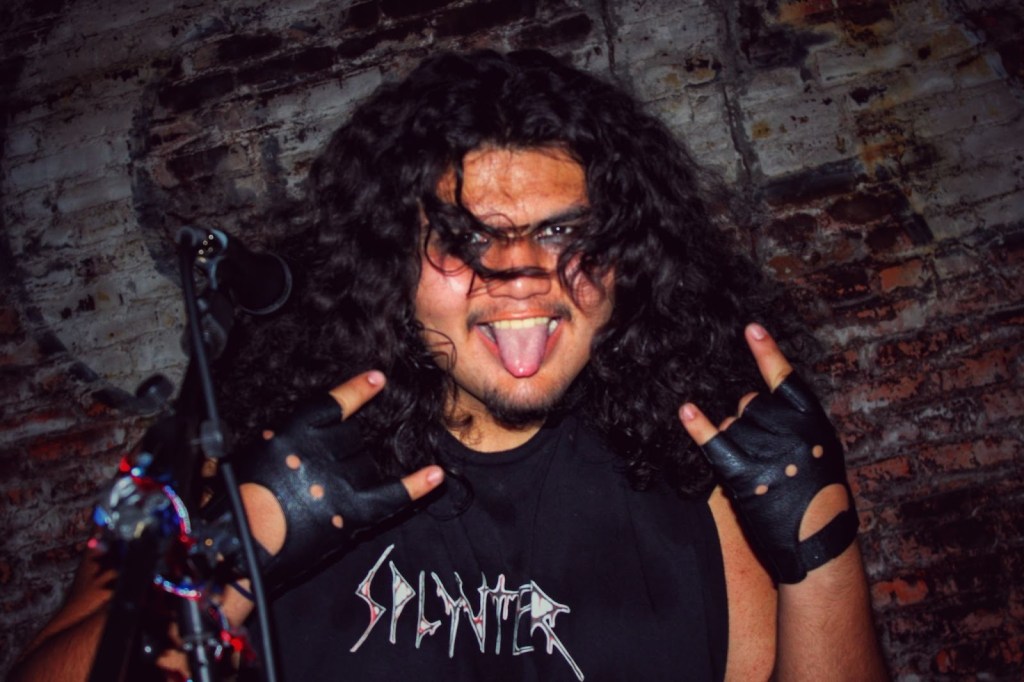 A member of the thrash metal band SPLYNTER performing energetically on stage, wearing a black tank top with the band's logo, showing the rock 'n' roll hand gesture.
