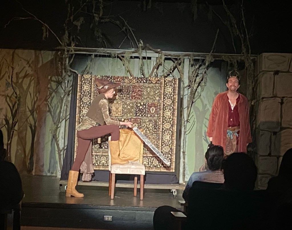 A theatrical performance scene featuring a female actor in costume wielding a sword while balancing on a chair, with a male actor standing beside her, both interacting in front of a decorative backdrop.