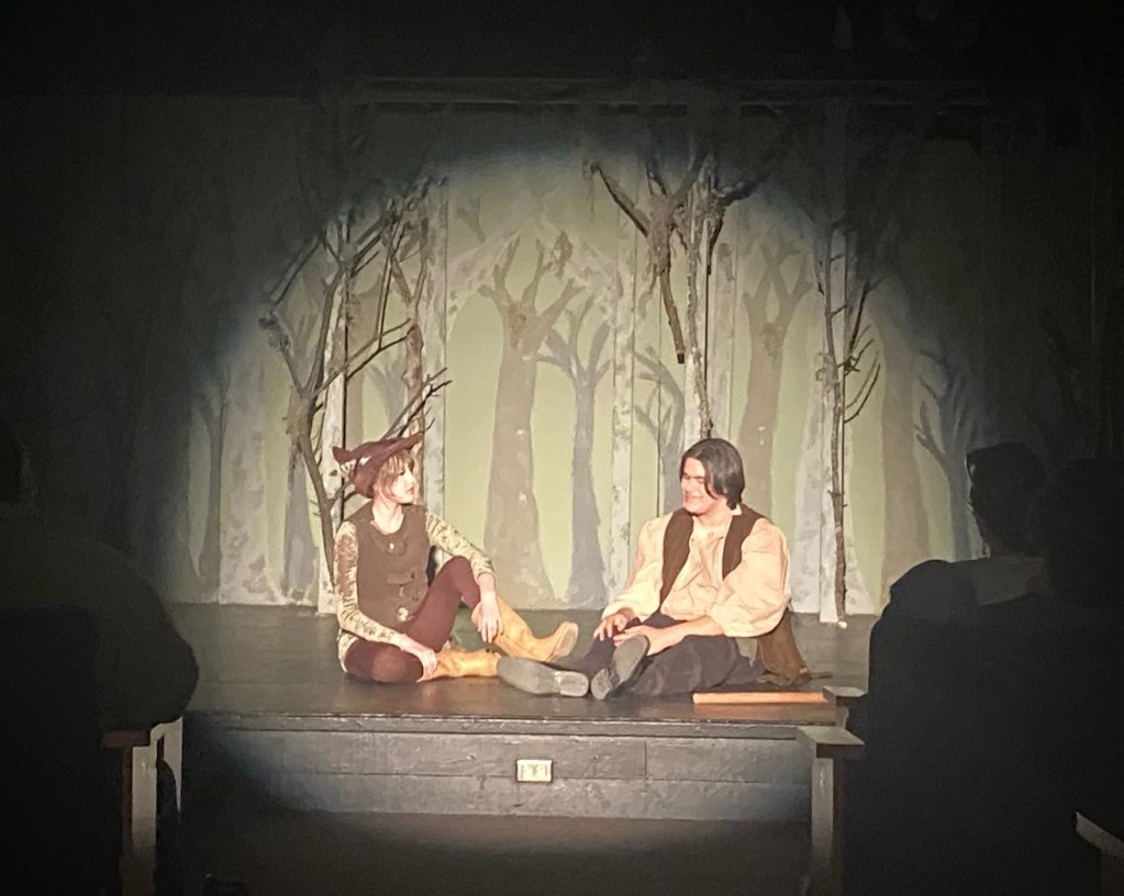 Two actors performing on stage in a wooded setting, one wearing a costume with horns and the other in a historical outfit, sitting on the floor with a warm spotlight highlighting them.