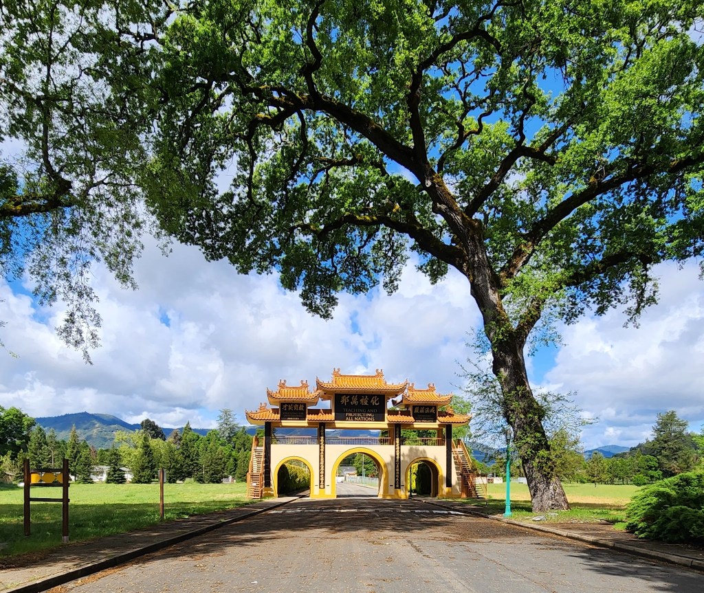 A wide shot of a traditional archway with a colorful roof, surrounded by lush greenery and a large oak tree, under a partly cloudy sky.