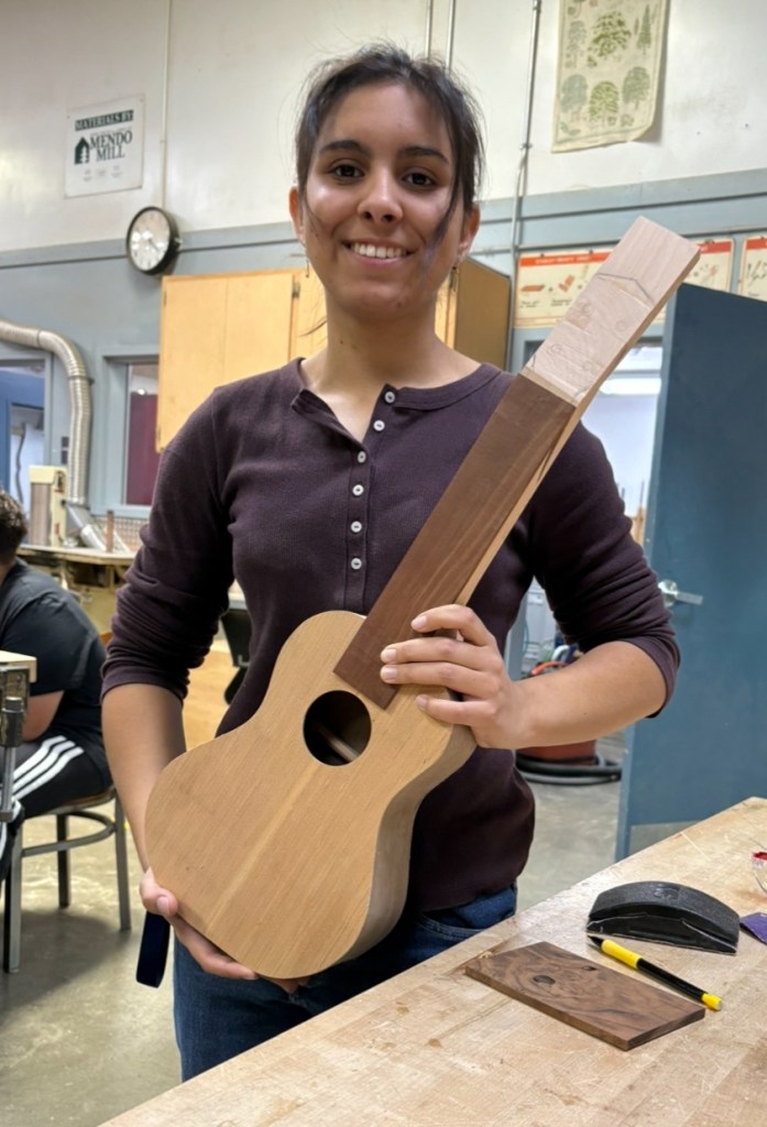 A young woman smiles while holding an unfinished ukulele made of wood in a workshop, with various tools and materials visible on the table.